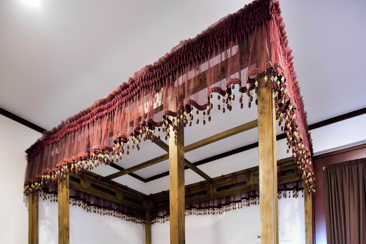 Una cama con dosel sobre cuatro postes, con un cojín rojo, estructura de madera y una ventana al pie de la cama.