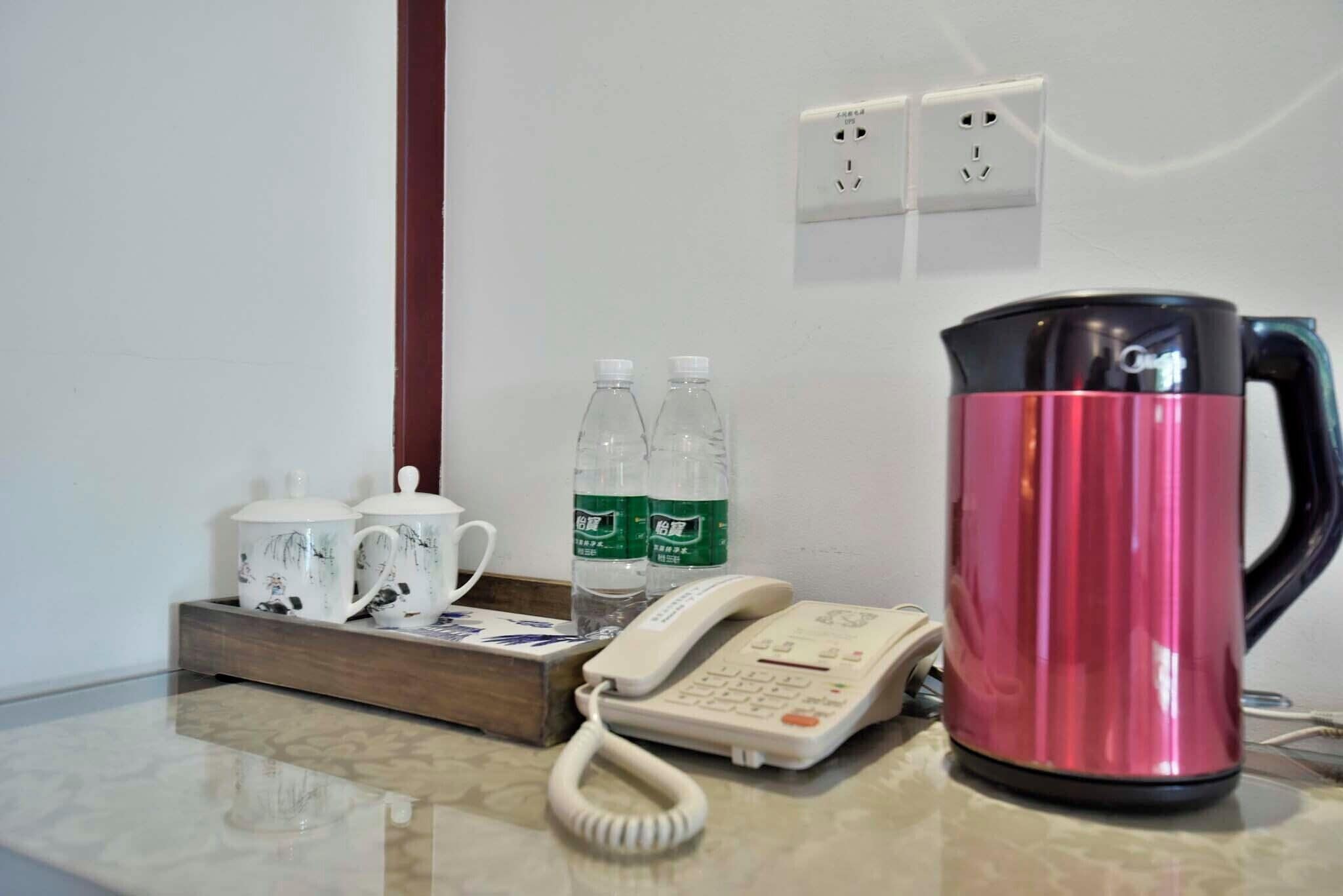 Une chambre d’hôtel avec un buffet en bois sur lequel sont posées deux théières, deux bouteilles d’eau et un téléphone. On peut également apercevoir une bouilloire électrique rose sur la table.