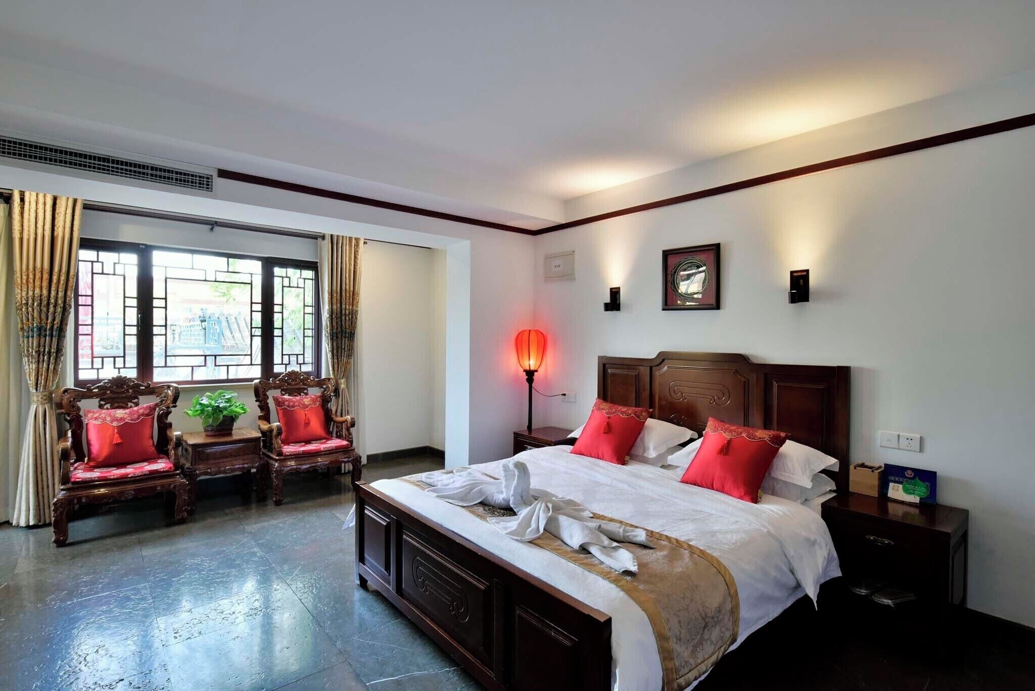 Habitación de hotel con una cama grande, dos sillas de madera, una mesita de noche, una lámpara y un ventanal con cortinas.