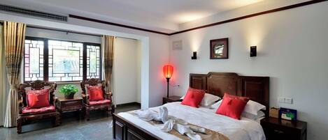 Habitación de hotel con una cama grande, dos sillas de madera, una mesita de noche, una lámpara y un ventanal con cortinas.