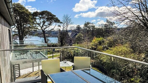 Een terras met glazen reling, rieten meubilair en uitzicht op een meer en bergen.