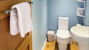 Een badkamer met een houten deur, een witte handdoek aan een metalen handdoekhouder, een wit toilet, een witte wastafel en een kleine prullenbak.
