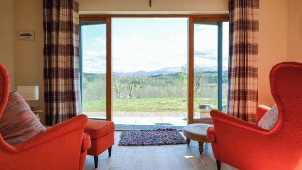 A room with two red armchairs, a small table, and a view of a grassy landscape through large glass doors. - 4 bedroom accommodation in Gairlochy, near Spean Bridge (Gairlochy, near Spean Bridge)
