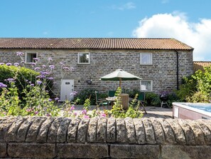 En hytte af sten med et tag af fliser, en dør med teksten 'Cottage' og en lille terrasse med bord, stole og parasol.