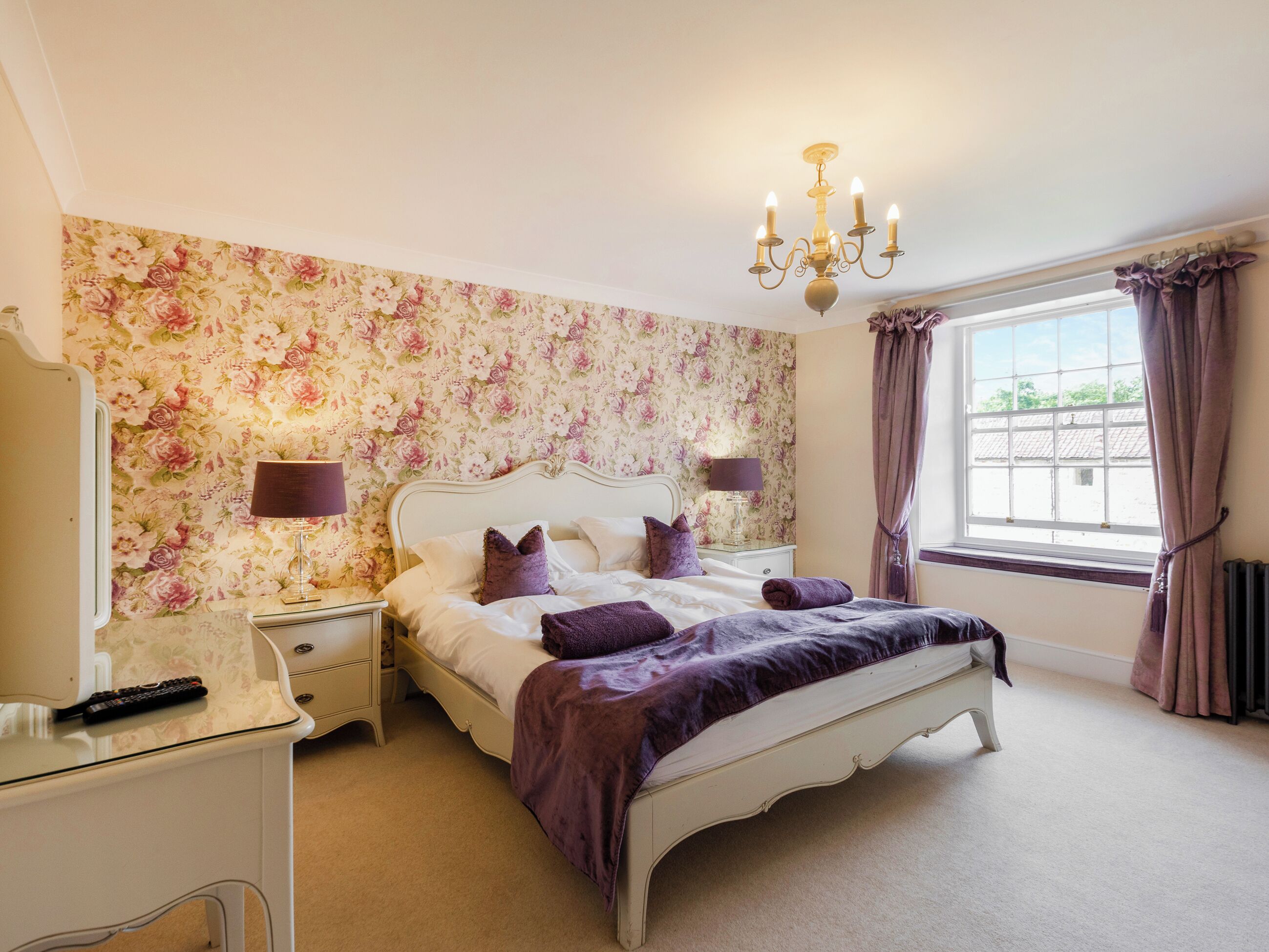 A bedroom with a floral wallpaper, a white bed, purple bedding, a chandelier, a nightstand, a lamp, a window with curtains, and a radiator.