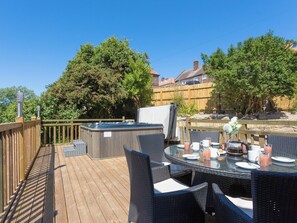 Eine Holzterrasse mit Whirlpool, Sitzgelegenheiten im Freien und Blick auf Bäume sowie ein Gebäude im Hintergrund.