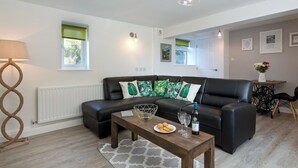Ett modernt vardagsrum med en svart lädersoffa, ett träbord och en lampa. Det finns fönster med gröna gardiner, ett element och ett litet bord med en vas med blommor.