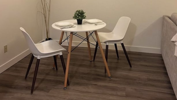 Uma pequena área de refeições com uma mesa redonda e duas cadeiras brancas, uma pequena planta em vaso e gramíneas decorativas altas contra uma parede lisa.