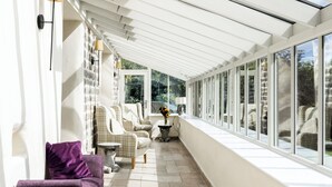 Eine sonnendurchflutete überdachte Terrasse mit Sitzgelegenheiten, einem Tisch und Blick auf Grünflächen.