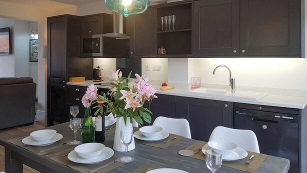A modern kitchen with dark cabinets, a white countertop, and a dining area set for six with white plates and glasses. - 3 bedroom accommodation in Whitby (Whitby)
