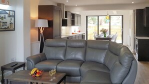 A modern living room with a grey leather sofa, a small table with fruit and wine glasses, a floor lamp, and a kitchen in the background. - 3 bedroom accommodation in Whitby (Whitby)