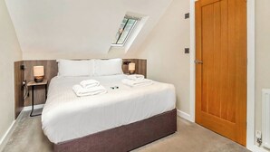 A neatly made bed with white linens, a wooden headboard, and a bedside table with a lamp. There is a skylight above the bed, and a wooden door to the right. - 4 Monkbar Mews - UK2693 (York)