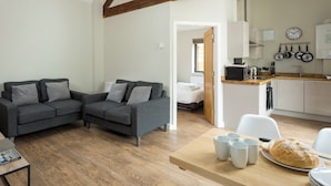 Een moderne leefruimte met een grijze bank, houten vloeren, een keuken met witte kastjes en een eethoek met een tafel gedekt voor twee personen.