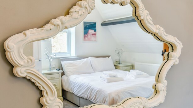 A bedroom with a large ornate mirror, a bed with white linens, a nightstand with a lamp, and a window with a view. - 2 bedroom accommodation in Whitby (Whitby)
