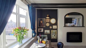 A dining area with a table set for two, a vase of flowers on the windowsill, and a wall decorated with framed pictures and a mirror. - A historical cottage, tucked away in the heart of Whitby’s old town. (Whitby)