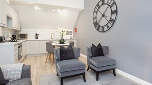 A modern kitchen with white cabinets, a microwave, and a sink. There are two grey armchairs and a dining table with chairs. A large clock is on the wall. - Minsters Reach Apartment Four - UK2749 (York)