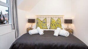 A hotel room with a large bed, two nightstands with lamps, a window with curtains, and a view of buildings outside. - Minsters Reach Apartment Four - UK2749 (York)