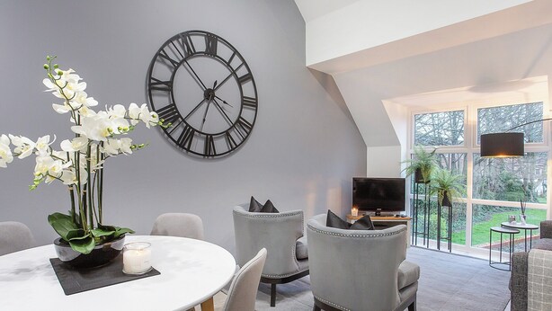 A modern living room with a large clock, a dining table, grey chairs, and a TV. - Minsters Reach Apartment Four - UK2749 (York)