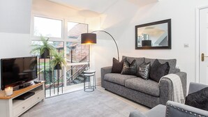 A living room with a TV, sofa, and a balcony with a view of a brick building. - Minsters Reach Apartment Four - UK2749 (York)