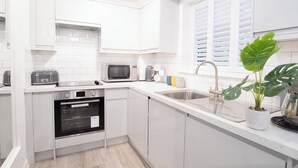 Et moderne køkken med hvide skabe, indbygget ovn, mikroovn, vask og et vindue med persienner.