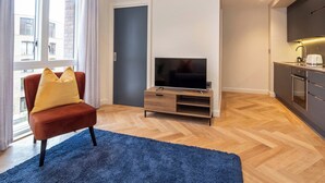Un apartamento moderno con una televisión sobre un mueble de madera, una silla roja con cojines amarillos, una cocina con fregadero y tostadora, y un ventanal con cortinas.