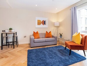 A modern living room with a grey sofa, orange cushions, a blue rug, and a small dining table with stools. - 1 bedroom accommodation in York (York)