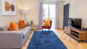 Una sala de estar moderna con un sofá gris, cojines naranjas, un tapete azul, un sill ón rojo, un televisor y un ventanal grande con cortinas.
