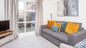 A living room with a grey sofa, a television on a wooden stand, and a large window with curtains. - Minsters Reach Apartment Two - UK2747 (York)