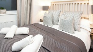 A neatly made bed with grey and white patterned pillows, a grey quilted bedspread, and rolled white towels on the bed. - Minsters Reach Apartment Two - UK2747 (York)
