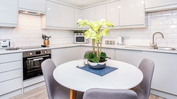 A modern kitchen with white cabinets, a built-in oven, a microwave, and a sink. There is a round table with chairs and a vase of yellow flowers. - Minsters Reach Apartment Two - UK2747 (York)
