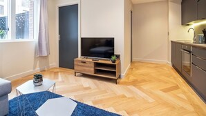 Un appartamento moderno con una TV su un supporto in legno, una cucina con pensili scuri e un'ampia finestra con tende.