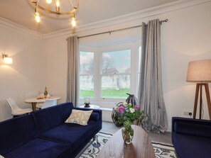 Ein Wohnzimmer mit einem blauen Sofa, einem kleinen Tisch mit Blumenvase, einer Lampe und Blick auf eine Steinsäule und Vegetation draußen.