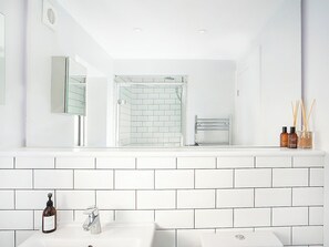 Ein modernes Badezimmer mit weißen, rechteckigen Fliesen, einer Glasdusche, einem weißen Waschbecken und einem Spiegel darüber. Auf der Ablage befinden sich zwei Seifenflaschen und ein Raumspray.