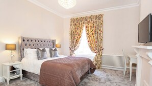 A bedroom with a tufted headboard bed, a brown bedspread, a nightstand with a lamp, a radiator, a television on a stand, and a window with floral curtains. - George Street Two - UK2767 (York)
