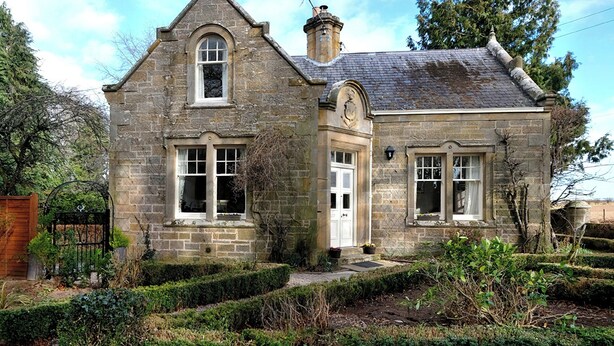 Une maison en pierre avec un toit à pignon, une entrée centrale et un petit porche agrémenté de plantes en pot.