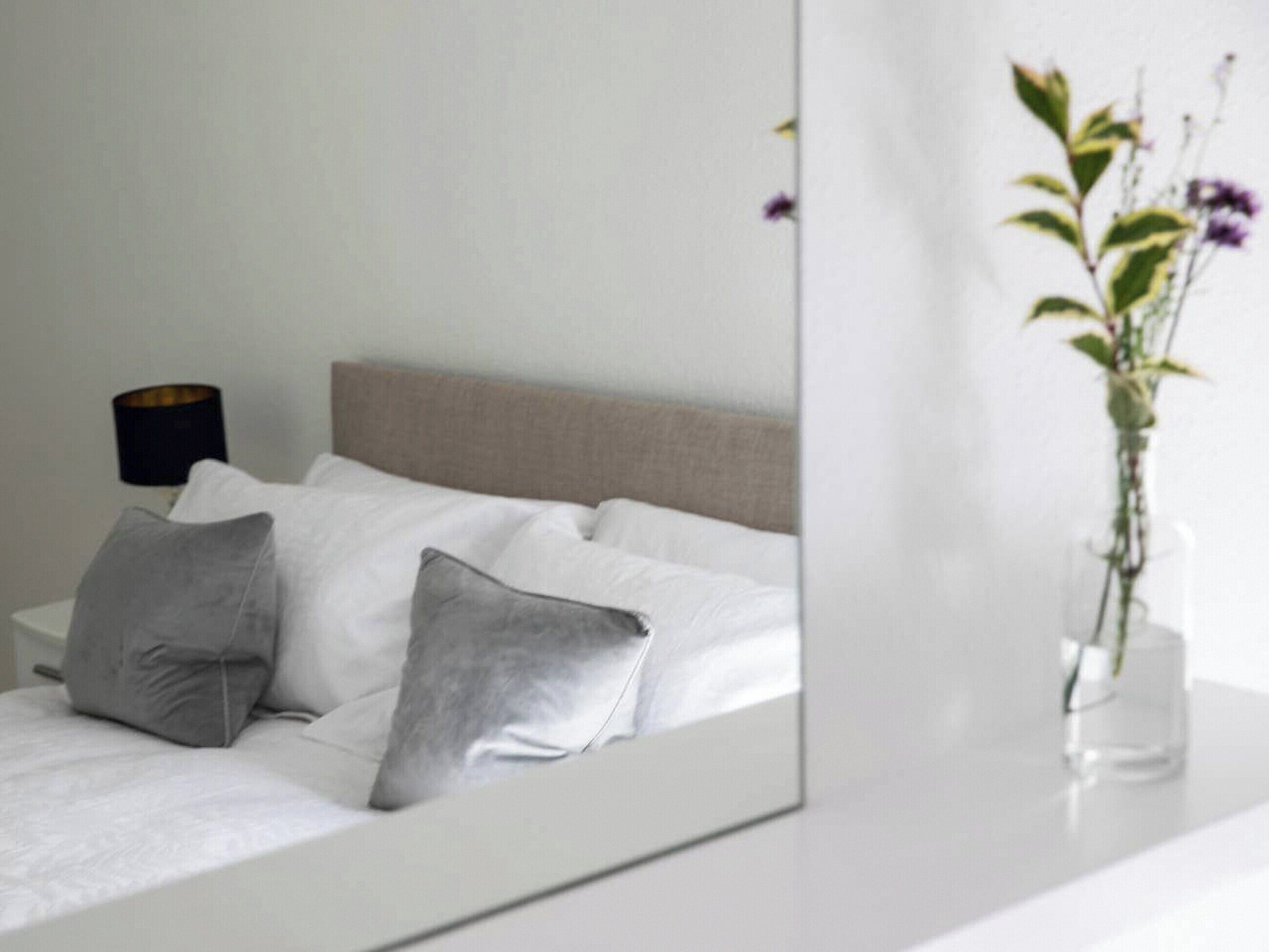 Un dormitorio con cama, almohadas, una mesita de noche, una lámpara y un florero con flores.