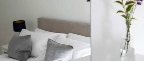Un dormitorio con cama, almohadas, una mesita de noche, una lámpara y un florero con flores.