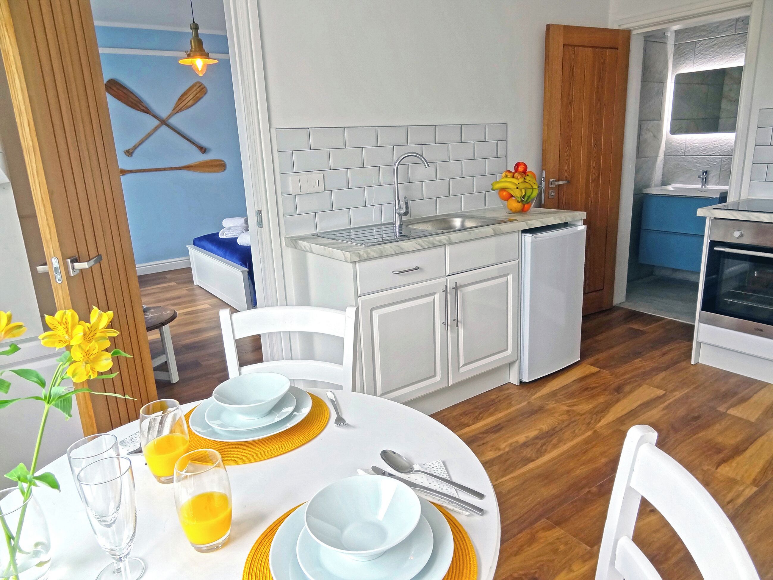 Cocina pequeña con gabinetes blancos, un lavabo y una estufa. Hay una mesa de comedor dispuesta con platos, vasos y cubiertos.