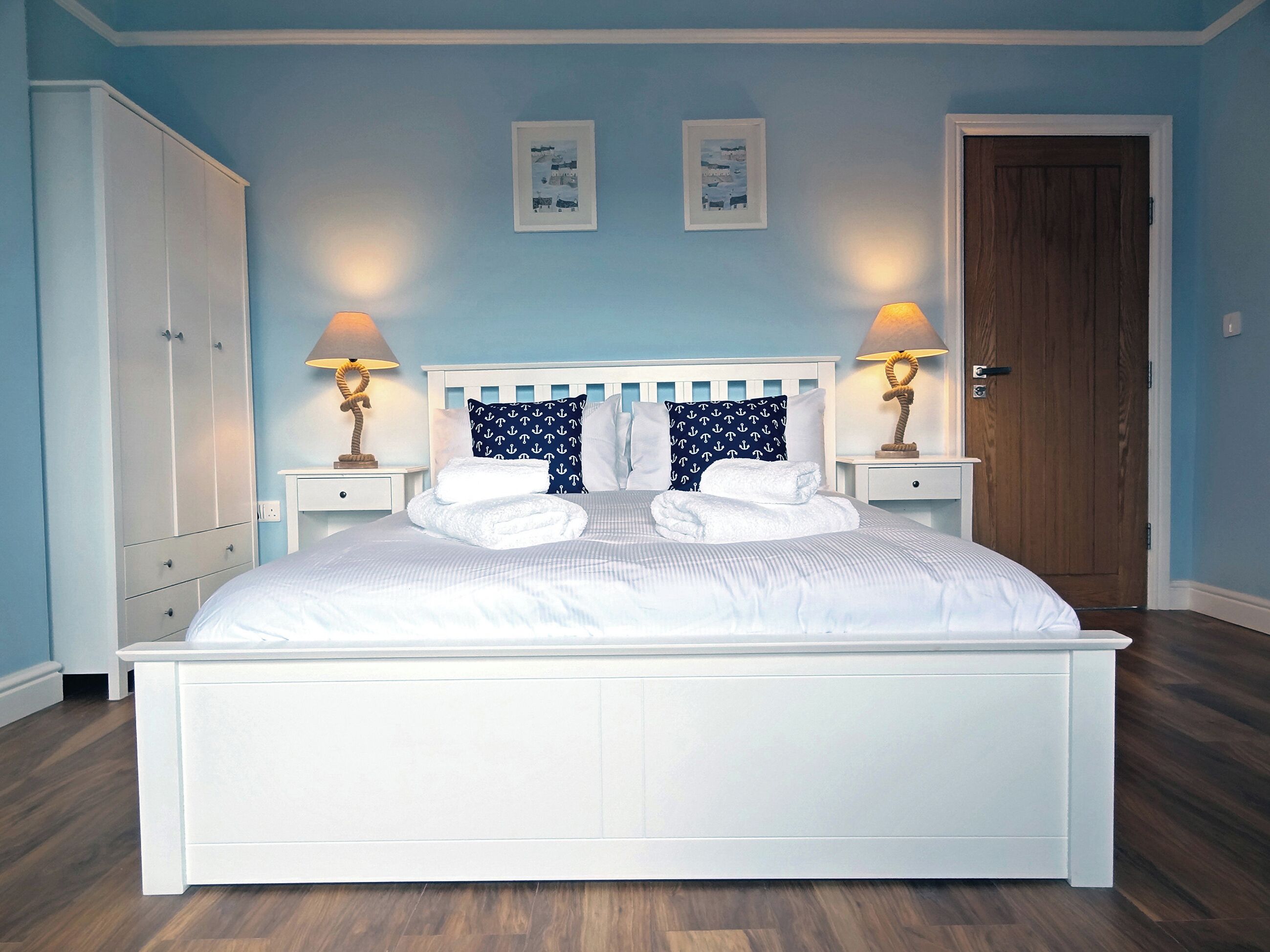 Una cama bien hecha con sábanas blancas y almohadas azul marino con estampados de anclas, flanqueada por dos mesitas de noche con lámparas, en una habitación con paredes azul claro y suelo de madera.