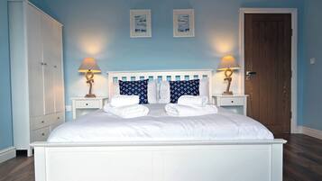 Una cama bien hecha con sábanas blancas y almohadas azul marino con estampados de anclas, flanqueada por dos mesitas de noche con lámparas, en una habitación con paredes azul claro y suelo de madera.