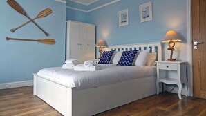 Un lit bien fait, avec du linge de lit et des serviettes blancs, un plancher en bois, une table de chevet blanche avec une lampe et une armoire.