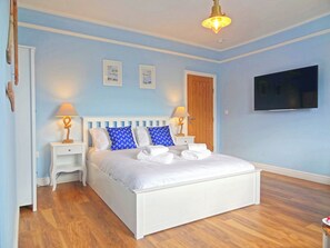Un dormitorio con una cama blanca, dos mesitas de noche con lámparas, una puerta de madera y un televisor montado en la pared.
