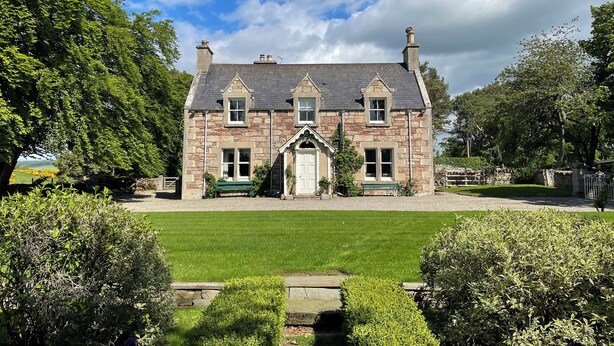 A stone house with a central entrance, surrounded by a well-maintained lawn and mature trees. - Balloan House-UK7383 (Dornoch)