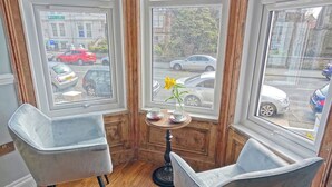 Ett hörnrum med två grå fåtöljer, ett litet träbord med en vas med gula blommor och utsikt över gatan genom stora fönster.