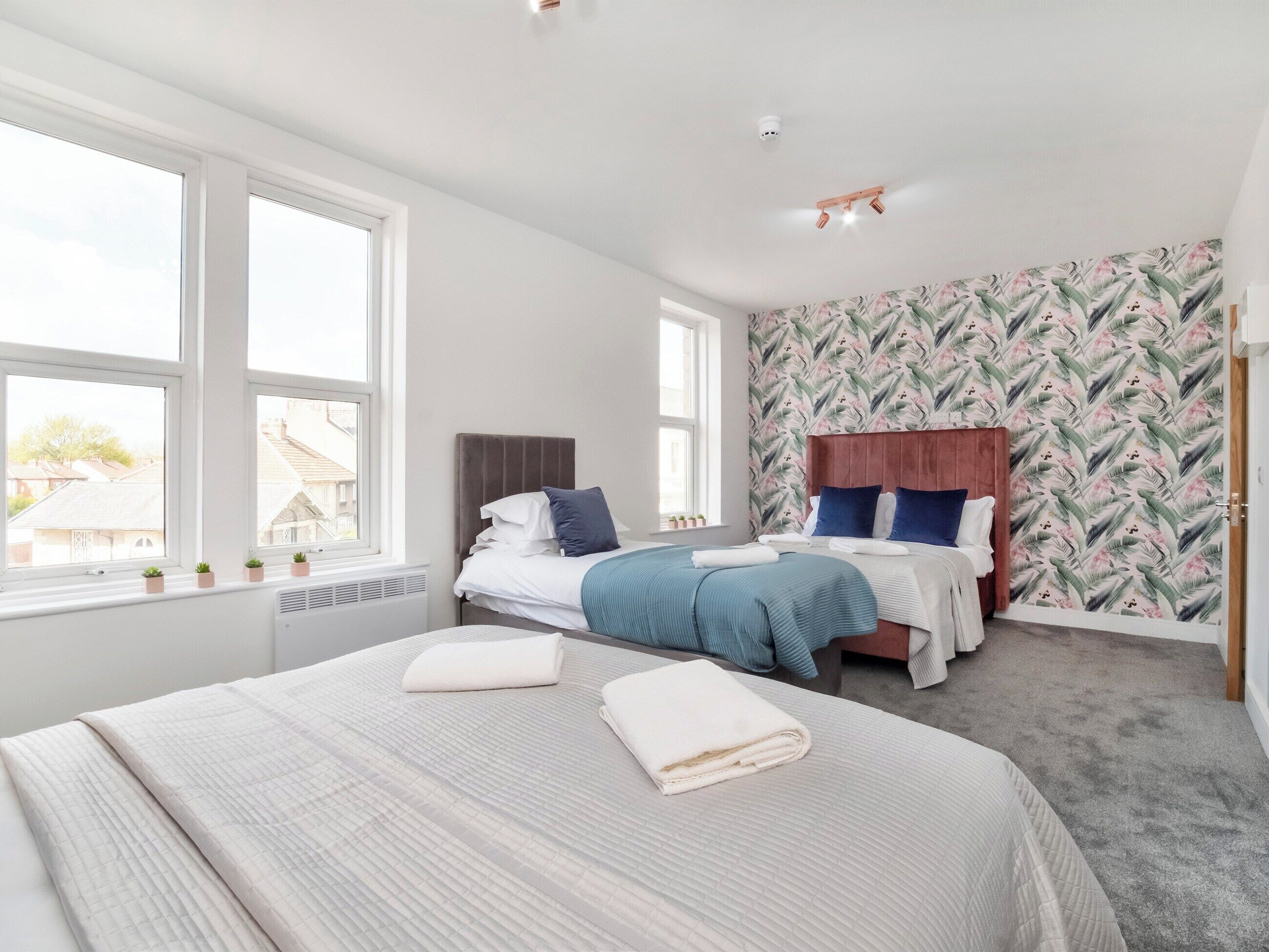 A bedroom with two beds, large windows, a radiator, and a floral wallpaper.
