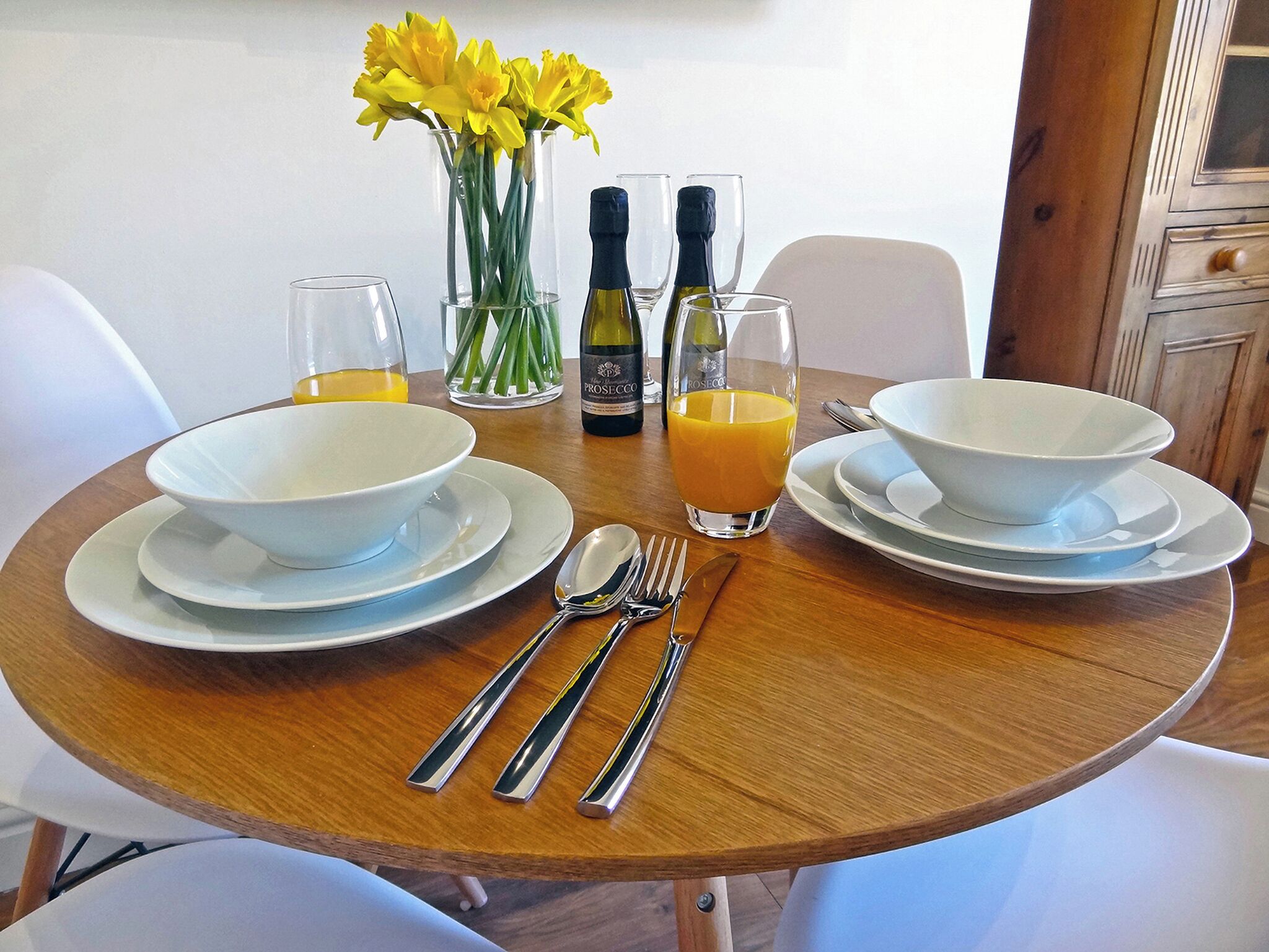 A dining table set with white bowls, plates, cutlery, and glasses, accompanied by a bottle of Prosecco and a glass of orange juice.