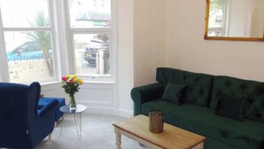 Een woonkamer met een groene bank, een blauwe fauteuil, een klein rond tafeltje en een bloemenvaas.