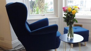 Een kamer met een blauwe fauteuil, een klein rond tafeltje met een bloevas, en uitzicht buiten door een raam.