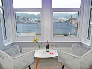 Habitación con dos ventanas amplias que ofrecen vista a un pueblo con edificios y colinas. Hay una mesa con una botella de champán, dos copas y un florero con flores.