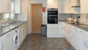 A modern kitchen with white cabinets, a built-in oven, a washing machine, and a trash bin. - Ty Fynnon Cottage-UK2786 (Penmaenmawr, near Conwy)